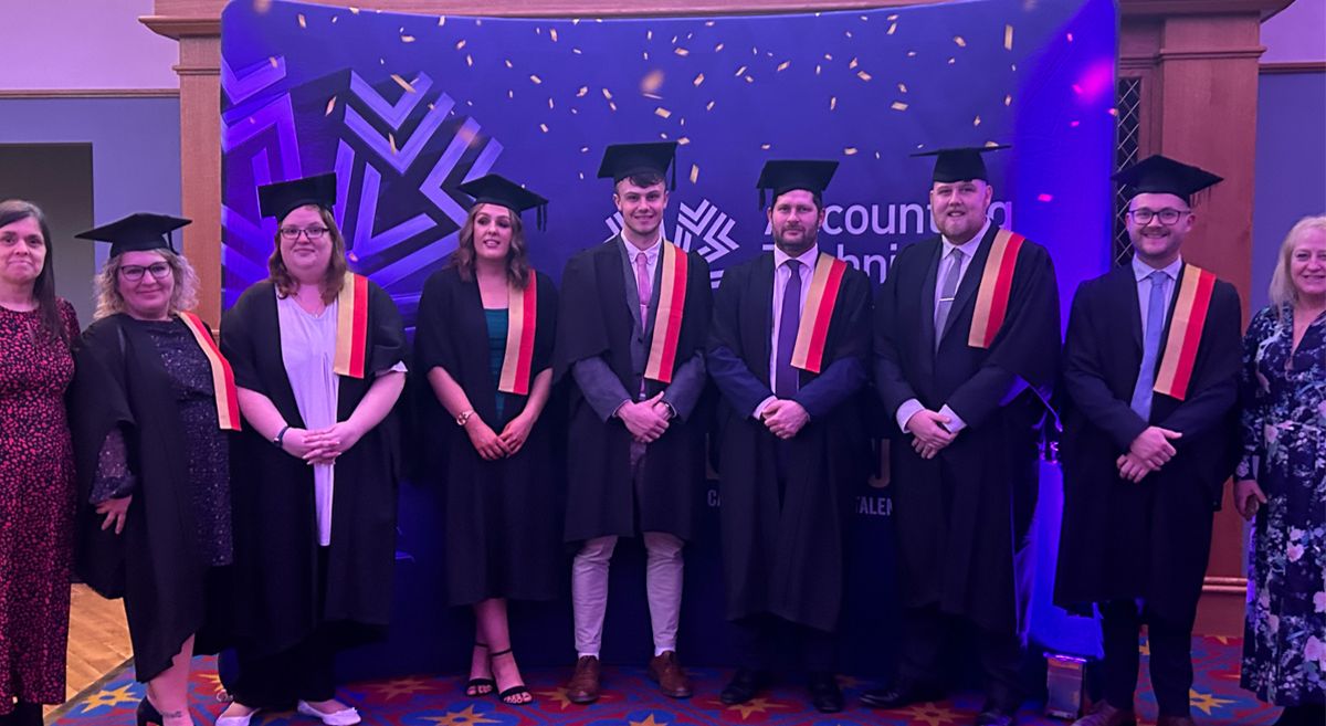 A line up of 9 people, 7 wearing graduation robe and cap
